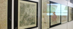 Oprawione mapy historyczne wiszą na białej ścianie. Widać fragmenty map terenowych i odbicie osoby.