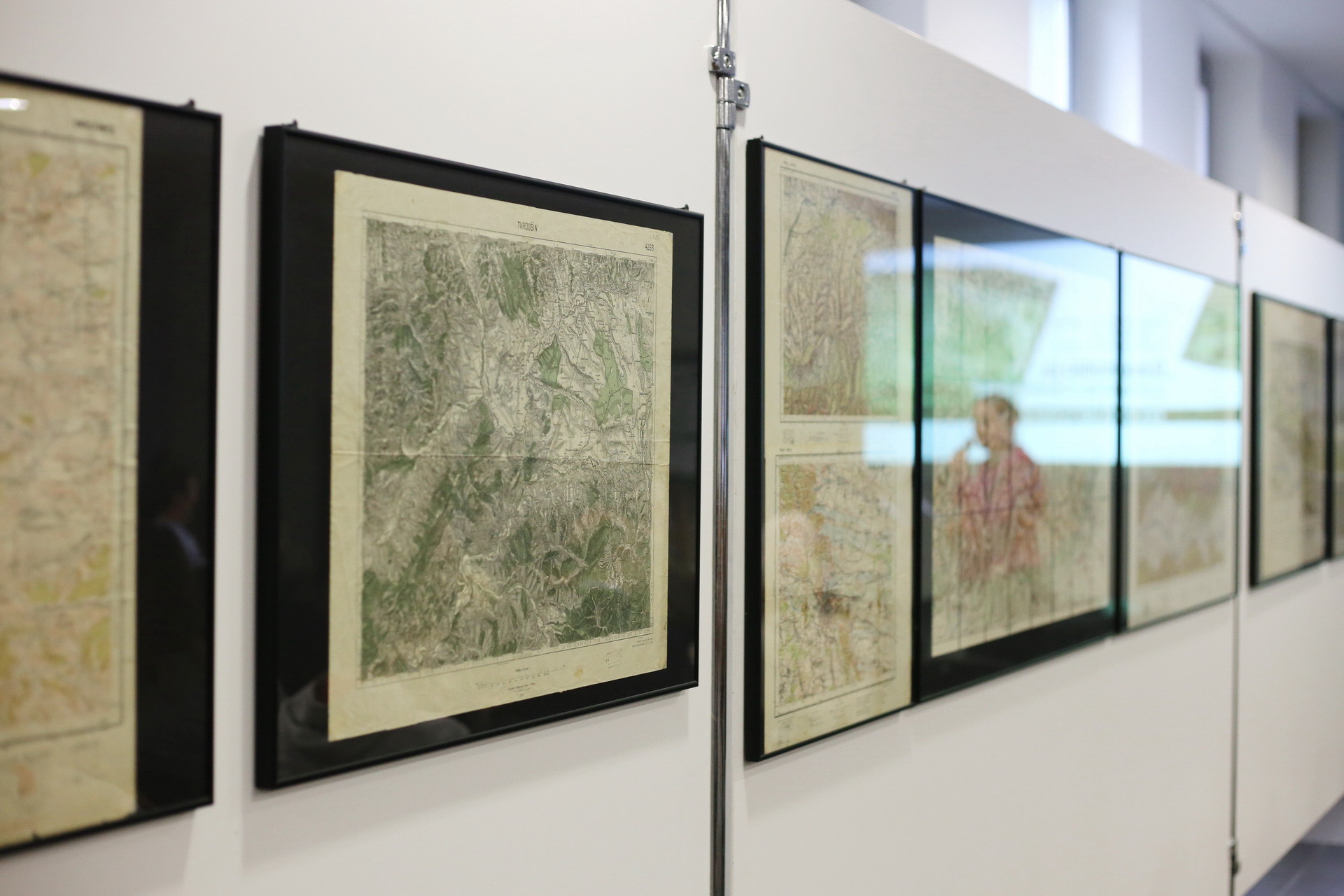 Oprawione mapy historyczne wiszą na białej ścianie. Widać fragmenty map terenowych i odbicie osoby.