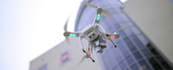 Biały dron z zielonymi i czerwonymi światłami unosi się przed nowoczesnym budynkiem. Kluczowe obiekty: dron, budynek.