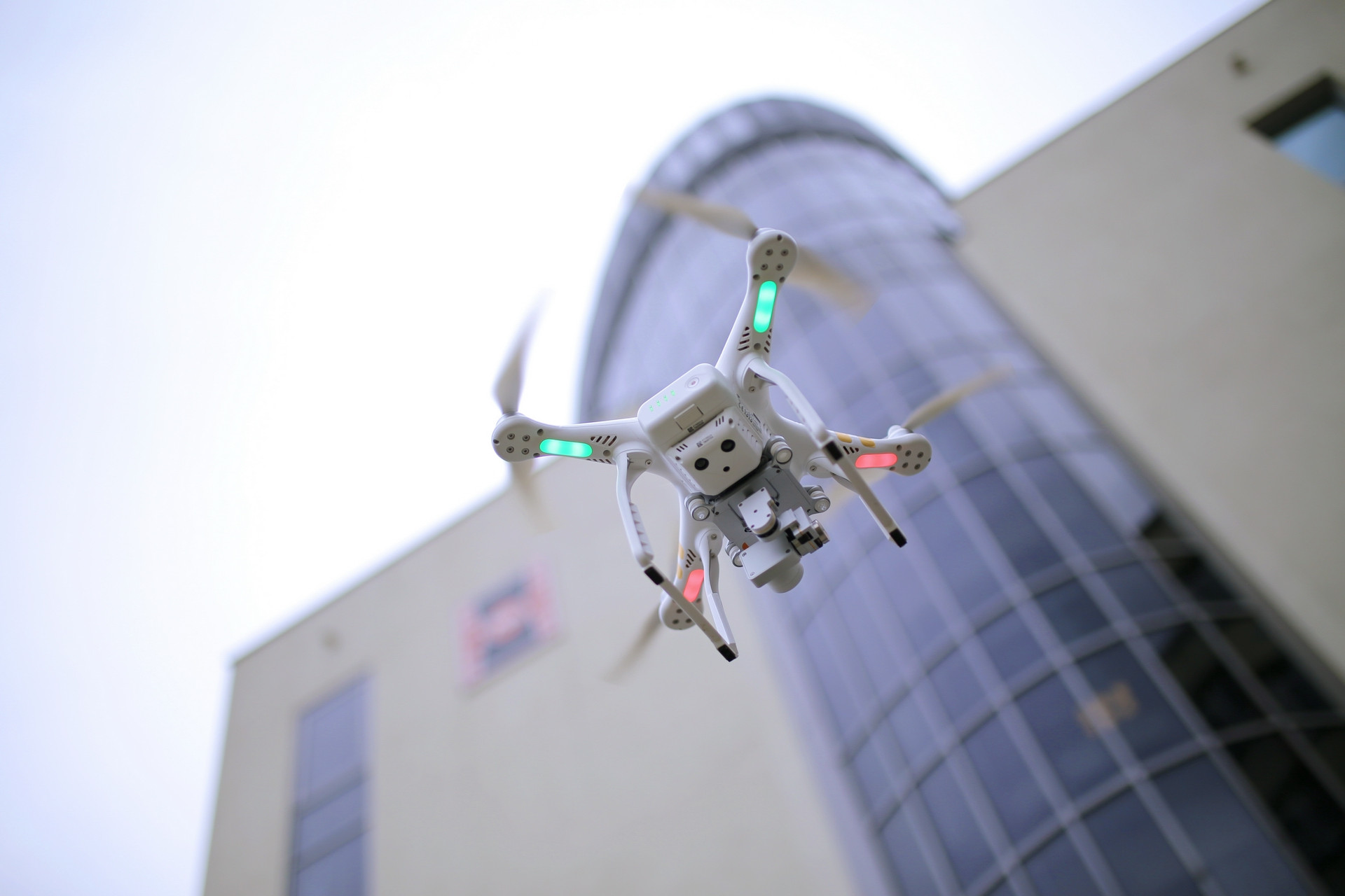Biały dron z zielonymi i czerwonymi światłami unosi się przed nowoczesnym budynkiem. Kluczowe obiekty: dron, budynek.